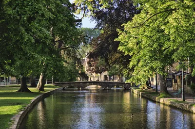 A scenic view of a river flowing through a village, with a stone arch bridge, green trees, and buildings on the banks.