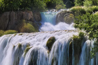 Multiple tiers of white waterfalls cascade over mossy rocks and lush green foliage in a sunny natural park.