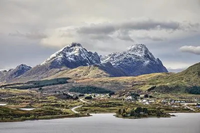 A landscape view of snow-capped mountains towering over a valley with a village, a winding road, and a calm lake. Overcast sky in Lofoten, Norway.