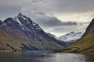 A wide shot of a Norwegian fjord with dark water, flanked by steep mountains with autumn trees and snowy peaks. A small cluster of red-roofed houses sits on a small island or peninsula.