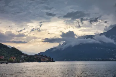 Landscape view of a village with a prominent tower on the shore of a lake, surrounded by mountains partially covered in clouds.
