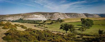A wide panoramic view of a landscape with rolling green hills, fields, and a prominent grey limestone escarpment under a clear blue sky with white clouds.
