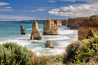 Coastal landscape featuring several large limestone sea stacks in turquoise ocean water, with waves crashing around their bases. Cliffs line the shore under a blue sky with white clouds.