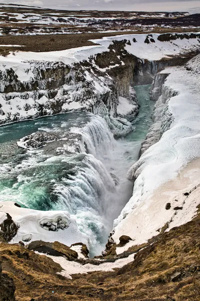 A powerful waterfall rushes through an icy, snow-covered gorge, surrounded by dark rock and open winter hills.