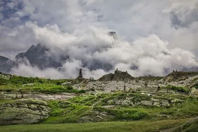 A landscape view of a mountain pass with many stacked stone cairns on green and rocky terrain. Thick white clouds obscure the peaks of large mountains in the background under a grey sky.