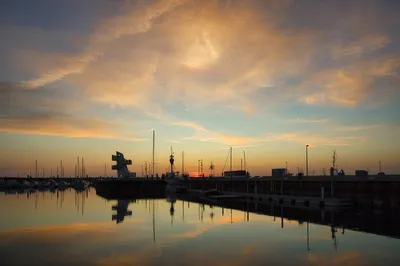 A wide shot of a marina at sunset with boats docked, a modern white sculpture on a pier, and colorful clouds reflected in the calm water.