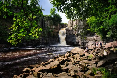 A powerful waterfall cascades into a rocky river, surrounded by cliffs and green trees, with people sitting on rocks in the foreground.