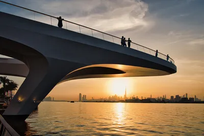 Silhouetted people on a modern bridge viewing platform overlooking Dubai Creek Harbour and the city skyline with the Burj Khalifa at sunset.