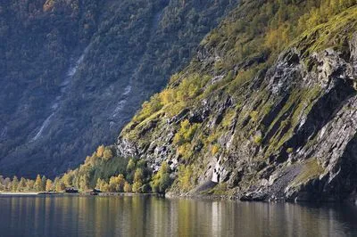 A wide shot of a Norwegian fjord with steep, rocky cliffs covered in autumn trees. Calm water reflects the sky and mountains, with small huts visible along the shoreline.
