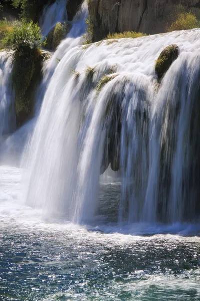 A long exposure photograph of a multi-tiered waterfall with white water cascading over mossy rocks into a turbulent pool below.