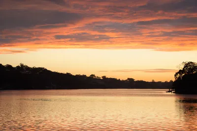 A sunrise scene over a calm river with orange and red clouds in the sky, reflecting on the water, and a dark silhouette of a tree-lined hill on the horizon.