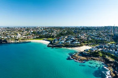 Aerial view of a coastal town with turquoise ocean, sandy beaches, and houses on hills under a clear blue sky.
