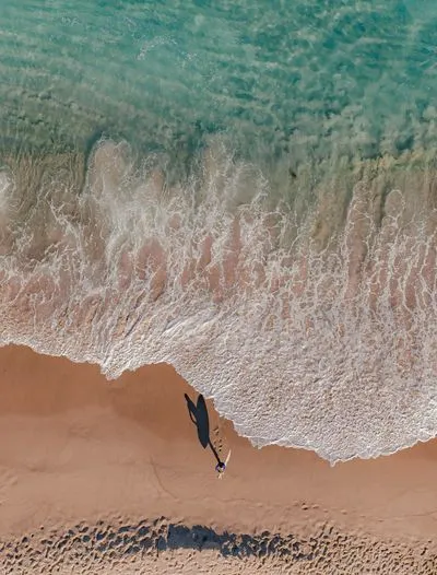 Aerial view of a person with a surfboard walking on a sandy beach next to turquoise water with breaking waves, casting a long shadow.