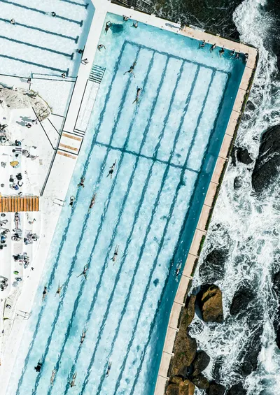 Aerial view of a large outdoor ocean swimming pool with people swimming and sunbathing, next to a rocky coastline with crashing waves.