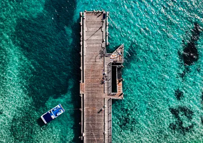 Aerial overhead shot of a long wooden jetty extending into clear turquoise ocean water. A white and blue boat is tied to the jetty. Several people are visible on the jetty, some sunbathing on a platfo