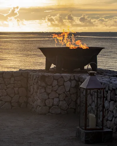 A large fire pit with flames burning brightly on a stone wall overlooking the ocean at sunset. A lantern with a candle sits in the foreground.