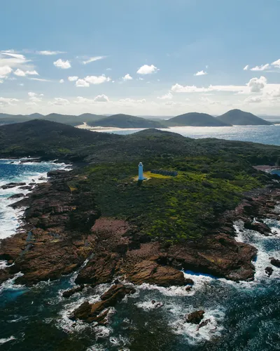 Aerial view of a white lighthouse on a green, rocky peninsula with waves crashing against the shore and hills in the background under a blue sky.