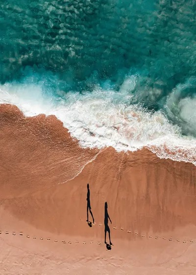 Aerial view of two long human shadows walking on a sandy beach next to turquoise ocean water with white waves.