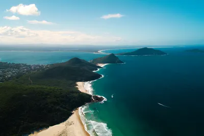 Aerial view of a coastal landscape with green hills, sandy beaches, and turquoise ocean water. Boats are visible in the bay.