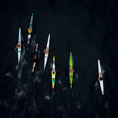 Aerial view of seven athletes in colorful surf skis paddling across dark water, leaving wakes behind them.