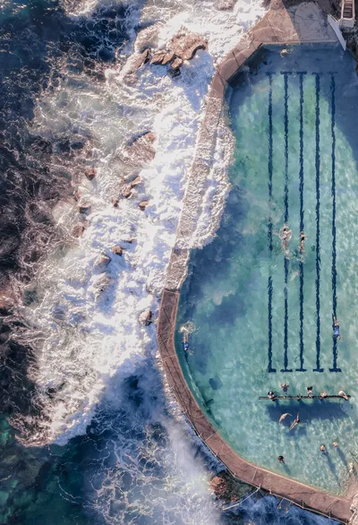 Aerial view of an ocean swimming pool with lane lines and people swimming, next to a rocky coastline with crashing waves.