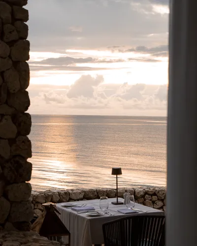 A set dinner table with a lamp, overlooking the ocean at sunset, framed by a stone wall and white pillar.