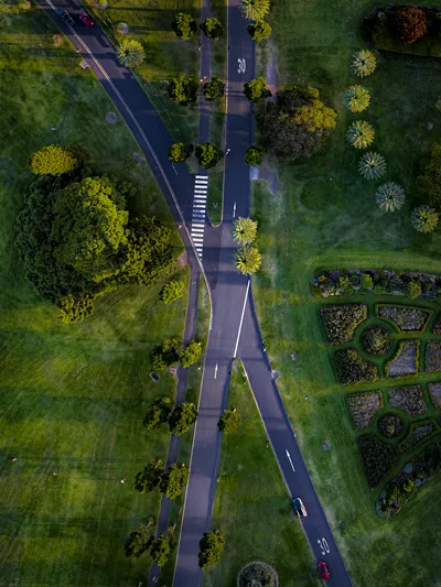 Aerial view of a park with winding roads, a pedestrian crossing, green lawns, trees, and geometrically patterned formal gardens.