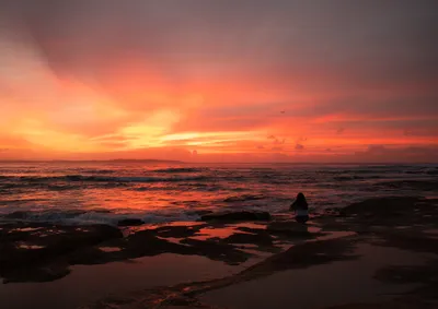 A person sits on dark rocks by the ocean, observing a vibrant sunrise with orange and red clouds, and waves breaking on the shore.