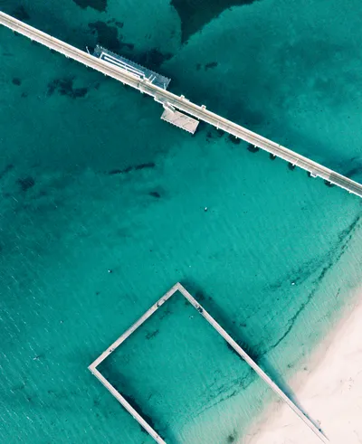 Aerial view of a long wooden jetty extending into clear turquoise ocean water, with a rectangular swimming enclosure near a sandy beach.