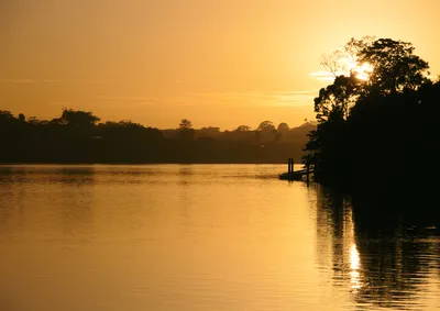 A golden sunrise over a calm river with silhouetted trees on the far bank and a small jetty with two people on the right.