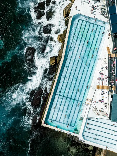 Aerial view of the Bondi Icebergs ocean pool with swimmers, adjacent to the ocean with waves crashing on rocks.