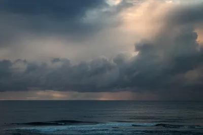 Ocean scene with dark, heavy clouds and a subtle light breaking through on the horizon, with waves and distant surfers.