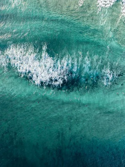 Aerial view of a vibrant turquoise ocean with many surfers on boards, some paddling, others near white breaking waves. The water shows varying shades of blue and green.