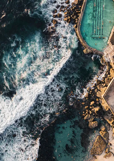 Aerial photograph of Bronte Baths, an ocean pool with swimmers, adjacent to breaking waves and a rocky shoreline.