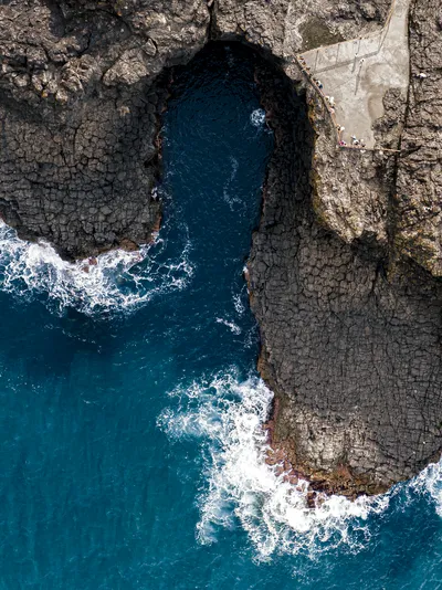 Aerial view of a rocky coastal inlet with dark blue water, a concrete viewing platform, and people.