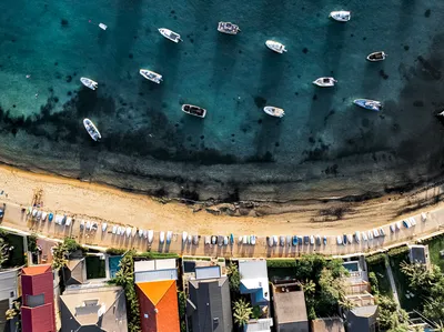 Aerial photograph of a bay with many boats anchored in turquoise water, a sandy beach, and houses along the shore.