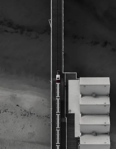 Aerial view of Busselton Jetty with a small train on tracks, next to white rectangular buildings, with beach and ocean on either side.