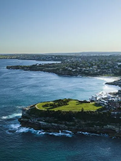 Aerial view of a green grassy headland with a winding path, rocky coastline, and a sandy beach. A city with numerous buildings stretches along the coast under a clear blue sky.